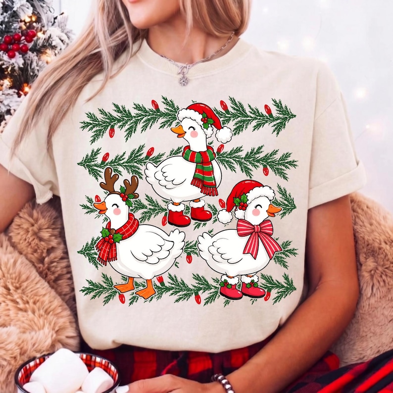 Może przedstawiać: Kremowy t-shirt z grafiką przedstawiającą trzy białe kaczki w świątecznych akcesoriach, takich jak czapki Mikołaja, szaliki i poroże renifera, na tle zielonych gałązek sosnowych z czerwonymi lampkami choinkowymi.