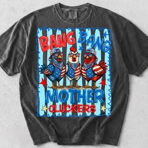 May include: A charcoal gray t-shirt featuring a graphic of three chickens wearing patriotic star-spangled bandanas and sunglasses. The text reads BANG BANG MOTHER CLUCKERS in red and blue lettering against a background of vertical blue stripes.