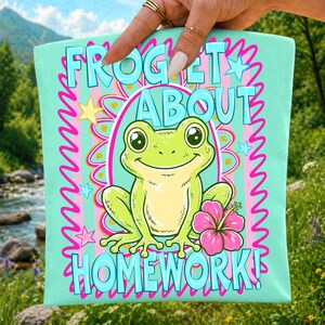 Pu&ograve; includere: Un quadrato di tessuto verde menta chiaro con un'illustrazione centrale di una rana verde sorridente. Il testo FROGET ABOUT HOMEWORK &egrave; stampato in azzurro. Il design include un fiore di ibisco rosa, stelle gialle e linee rosa.