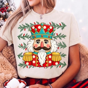 Puede incluir: Una camiseta de color crema con el gr&aacute;fico de un cascanueces que lleva una corona roja y azul verdoso, una chaqueta roja con charreteras doradas y una barba blanca, rodeado por un patr&oacute;n de ramas de pino verdes y peque&ntilde;as luces navide&ntilde;as rojas.