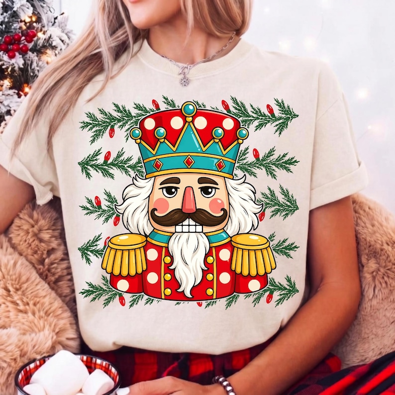 Puede incluir: Una camiseta de color crema con el gr&aacute;fico de un cascanueces que lleva una corona roja y azul verdoso, una chaqueta roja con charreteras doradas y una barba blanca, rodeado por un patr&oacute;n de ramas de pino verdes y peque&ntilde;as luces navide&ntilde;as rojas.