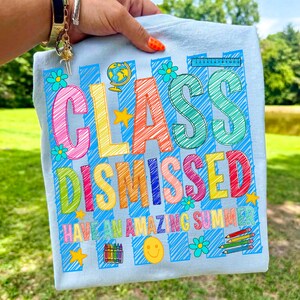 May include: A light blue t-shirt featuring a colorful graphic with the text "CLASS DISMISSED" and "HAVE AN AMAZING SUMMER". The design includes scribbled letters, a globe, a ruler, a smiley face, crayons, and flowers in bright colors.