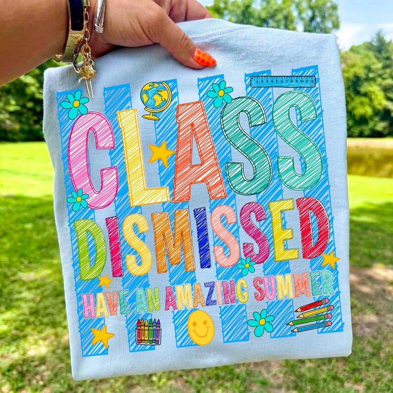 May include: A light blue t-shirt featuring a colorful graphic with the text "CLASS DISMISSED" and "HAVE AN AMAZING SUMMER". The design includes scribbled letters, a globe, a ruler, a smiley face, crayons, and flowers in bright colors.