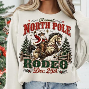 May include: A cream-colored crewneck sweatshirt featuring a graphic of Santa Claus riding a horse while holding a lasso. The text reads Annual NORTH POLE RODEO Dec. 25th. The design includes pine trees, a wooden fence, and red and green accents.
