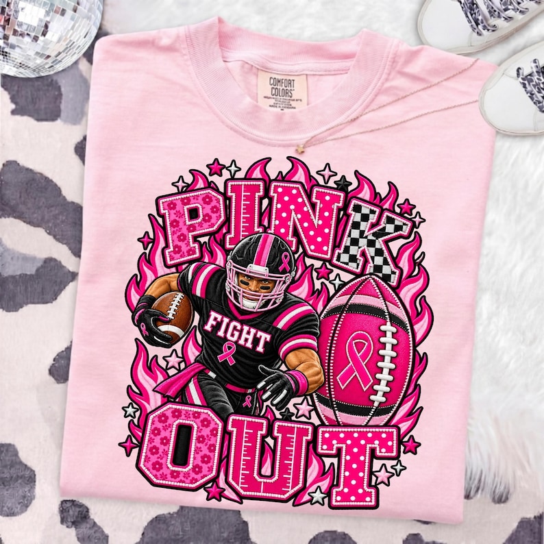 K&ouml;nnte beinhalten: Ein hellrosa T-Shirt mit einer Grafik eines Footballspielers in schwarzer Uniform mit rosa Akzenten. Der Text lautet PINK OUT und FIGHT. Ein rosa Football mit einem Schleifensymbol ist abgebildet, umgeben von rosa Flammen und Sternen.