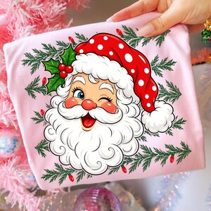 May include: A light pink t-shirt featuring a graphic illustration of a winking Santa Claus wearing a red hat with white polka dots. The design is surrounded by green pine branches, red holly berries, and small red Christmas light bulbs.