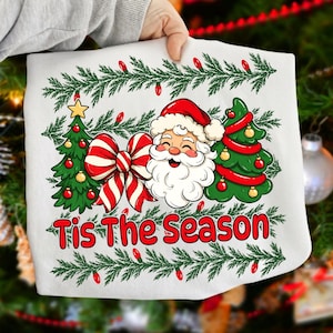 Puede incluir: Una sudadera blanca con un gr&aacute;fico de Santa Claus, dos &aacute;rboles de Navidad decorados y un gran lazo a rayas rojas y blancas. El texto "Tis The Season" est&aacute; impreso en rojo debajo del dise&ntilde;o, enmarcado por una guirnalda de pino verde con luces rojas.