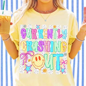 Pu&ograve; includere: Una t-shirt giallo pallido con una stampa grafica che riporta il testo "CURRENTLY CRASHING OUT" in lettere colorate, a scacchi e a tinta unita. Il design include una faccina sorridente che si scioglie, fiocchi rosa, stelle blu e piccoli fiori.
