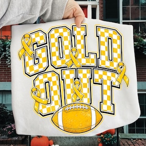 Puede incluir: Una camiseta blanca con el texto GOLD OUT en letras grandes de bloque con un patr&oacute;n de cuadros amarillos y blancos. El dise&ntilde;o incluye cuatro lazos amarillos de concienciaci&oacute;n y un gr&aacute;fico de un bal&oacute;n de f&uacute;tbol americano amarillo.