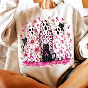 May include: A cream-colored sweatshirt featuring a graphic print of three ghosts and three black cats. The ghosts are decorated with pink floral patterns, leopard spots, and pink breast cancer awareness ribbons, surrounded by pink flowers and stars.