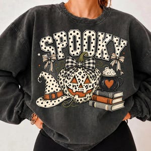 May include: A charcoal grey crewneck sweatshirt featuring a graphic print with the word SPOOKY in white polka-dot letters. The design includes a leopard-print jack-o'-lantern with a plaid bow, a polka-dot witch hat, a cauldron, and three books.