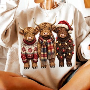 May include: A cream-colored sweatshirt featuring a graphic of three Highland cows. The left cow wears a red and white snowflake sweater, the middle cow wears a plaid scarf, and the right cow wears a Santa hat and a string of colorful Christmas lights.