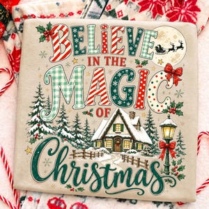 May include: A beige sweatshirt featuring the text "BELIEVE IN THE MAGIC OF CHRISTMAS" in colorful, patterned lettering. The design includes a snowy cabin, pine trees, a street lamp with a red bow, and a silhouette of Santa in a sleigh.