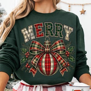 Puede incluir: Una sudadera verde oscuro de cuello redondo con la palabra MERRY en una fuente de cuadros sobre un gr&aacute;fico de adorno navide&ntilde;o. El adorno est&aacute; decorado con un lazo de cuadros rojo, verde y dorado, agujas de pino y bayas de acebo rojas.