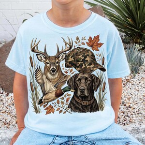 May include: A light blue t-shirt featuring a graphic print of a deer, a flying mallard duck, a black Labrador retriever wearing a collar, and a camouflage baseball cap, all surrounded by cattails and autumn leaves.