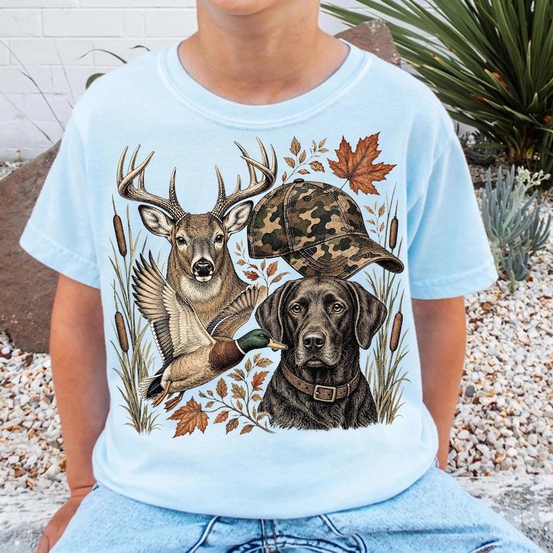 May include: A light blue t-shirt featuring a graphic print of a deer, a flying mallard duck, a black Labrador retriever wearing a collar, and a camouflage baseball cap, all surrounded by cattails and autumn leaves.