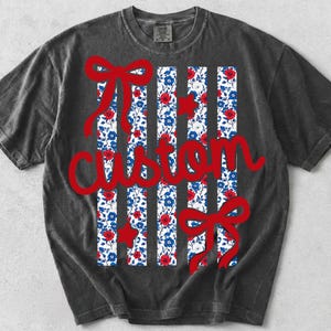 May include: A charcoal gray short-sleeve t-shirt featuring a graphic design of vertical stripes with a red, white, and blue floral pattern. The design includes two red bows, two red stars, and the word custom written in red cursive script.