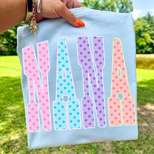 May include: A light blue t-shirt with the word "NANA" in large, colorful letters. The letters are outlined in white and filled with polka dots in pink, blue, purple, and peach. The shirt is held up by a hand.