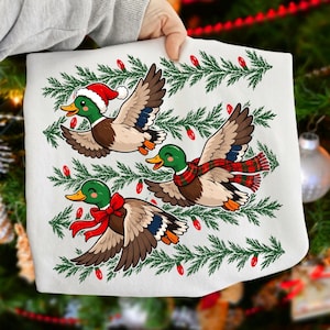 May include: A white t-shirt featuring a graphic print of three mallard ducks in flight. One duck wears a red Santa hat, one wears a red and green plaid scarf, and one wears a red bow. The ducks are arranged among horizontal pine branches with red lights.