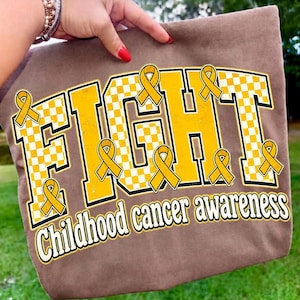 May include: A brown tote bag featuring a large graphic with the word FIGHT in bold yellow and white checkered block letters. Multiple yellow awareness ribbons are placed around the text, with the words childhood cancer awareness below.