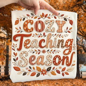 May include: A white t-shirt featuring the text Cozy Teaching Season in a decorative font. The letters are filled with orange, brown, and white patterns including gingham and floral prints. The design is surrounded by a border of autumn leaves.