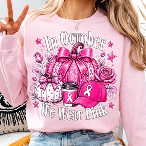 May include: A light pink crewneck sweatshirt featuring a graphic print of a large pink pumpkin with a bow, a smaller white polka-dot pumpkin, a coffee cup, and a baseball cap, all adorned with pink ribbons. The text reads In October We Wear Pink.
