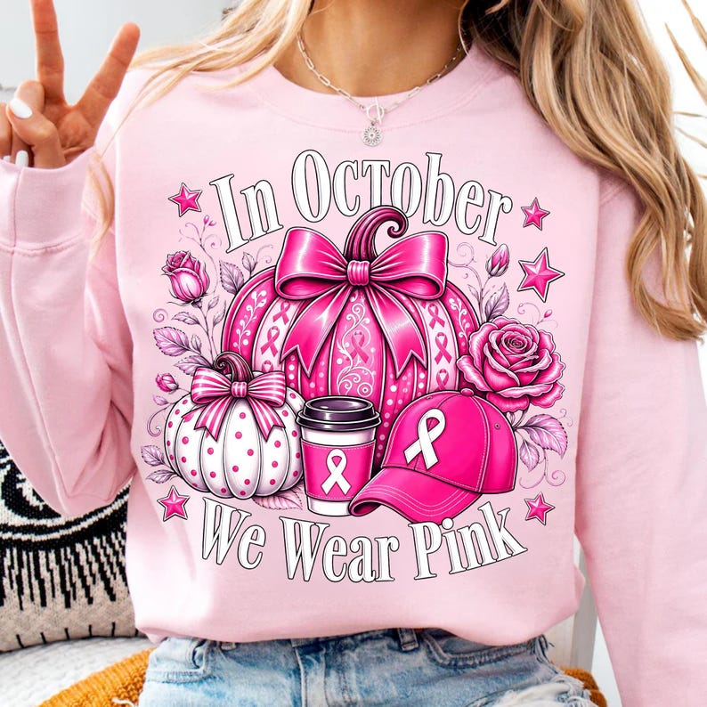 May include: A light pink crewneck sweatshirt featuring a graphic print of a large pink pumpkin with a bow, a smaller white polka-dot pumpkin, a coffee cup, and a baseball cap, all adorned with pink ribbons. The text reads In October We Wear Pink.