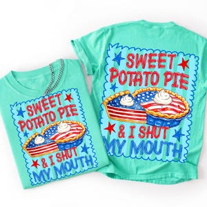 Op de afbeelding: Een mintgroen t-shirt met een afbeelding van een taart in het rood, wit en blauw van de Amerikaanse vlag, gegarneerd met slagroom. De tekst luidt SWEET POTATO PIE & I SHUT MY MOUTH, omringd door rode en blauwe sterren op een gestreepte achtergrond.
