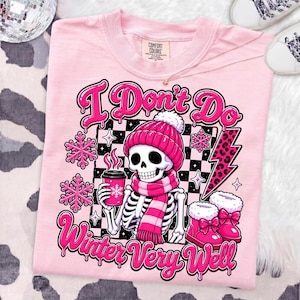 May include: A light pink t-shirt featuring a graphic of a skeleton wearing a pink beanie and scarf, holding a coffee cup. The design includes pink snowflakes, a leopard print lightning bolt, and the text I Don't Do Winter Very Well.