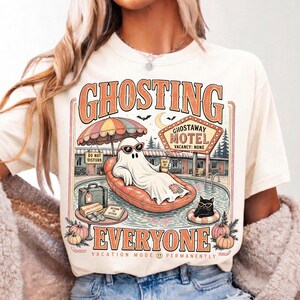 Puede incluir: Una camiseta de color crema con un gr&aacute;fico de un fantasma descansando en un flotador de piscina en el Ghostaway Motel. El dise&ntilde;o incluye el texto GHOSTING EVERYONE, VACATION MODE PERMANENTLY y OUT OF OFFICE, junto a un gato negro y calabazas.