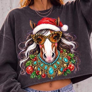 May include: A charcoal gray cropped sweatshirt featuring a graphic illustration of a horse wearing a red and white Santa hat, leopard print cat-eye glasses, and a decorative necklace with turquoise stones, gold bells, red ornaments, and holly.
