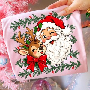 May include: A light pink t-shirt featuring a graphic illustration of Santa Claus and a reindeer. Santa wears a red hat and has a white beard. The reindeer has brown fur and a large red bow. Green pine branches and red lights frame the design.