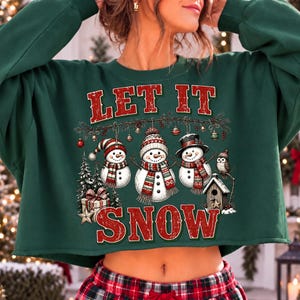Pu&ograve; includere: Una felpa corta verde scuro con una stampa grafica di tre pupazzi di neve che indossano cappelli e sciarpe, con la scritta LET IT SNOW in rosso. Il disegno include un abete, un regalo, una casetta per uccelli e un gufo sotto un ramo.