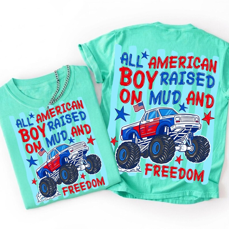 May include: A mint green t-shirt featuring a graphic of a red, white, and blue monster truck with an American flag. The text reads: ALL AMERICAN BOY RAISED ON MUD AND FREEDOM. The design includes blue and red stars on a light blue striped background.