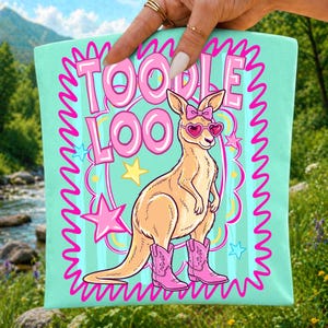 May include: A light mint green fabric item featuring a graphic of a tan kangaroo wearing pink heart-shaped sunglasses and pink cowboy boots. The kangaroo has a pink bow on its head. The text TOODLE LOO is printed in large pink letters above.