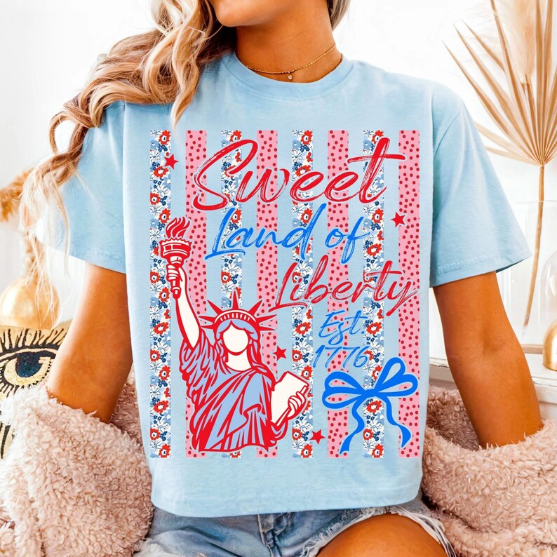 May include: Light blue t-shirt with a graphic featuring the Statue of Liberty holding a torch, with the text "Sweet Land of Liberty Est. 1776". The design includes red, white, and blue elements, including stripes, stars, and floral patterns.