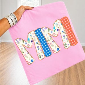 May include: A square light pink pillow cover featuring the word MIMI in large, bold letters. Each letter has a distinct pattern: floral prints with small colorful blossoms, a blue background with white stars, and a red background with white dots.