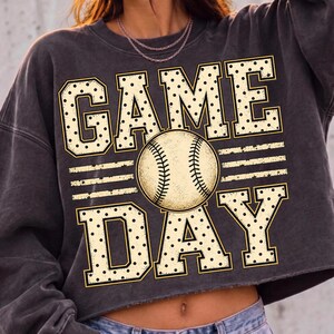 May include: A charcoal grey cropped sweatshirt featuring a graphic print with the text GAME DAY in large, cream-coloured block letters with black polka dots. A central baseball illustration with visible stitching sits between the two words.