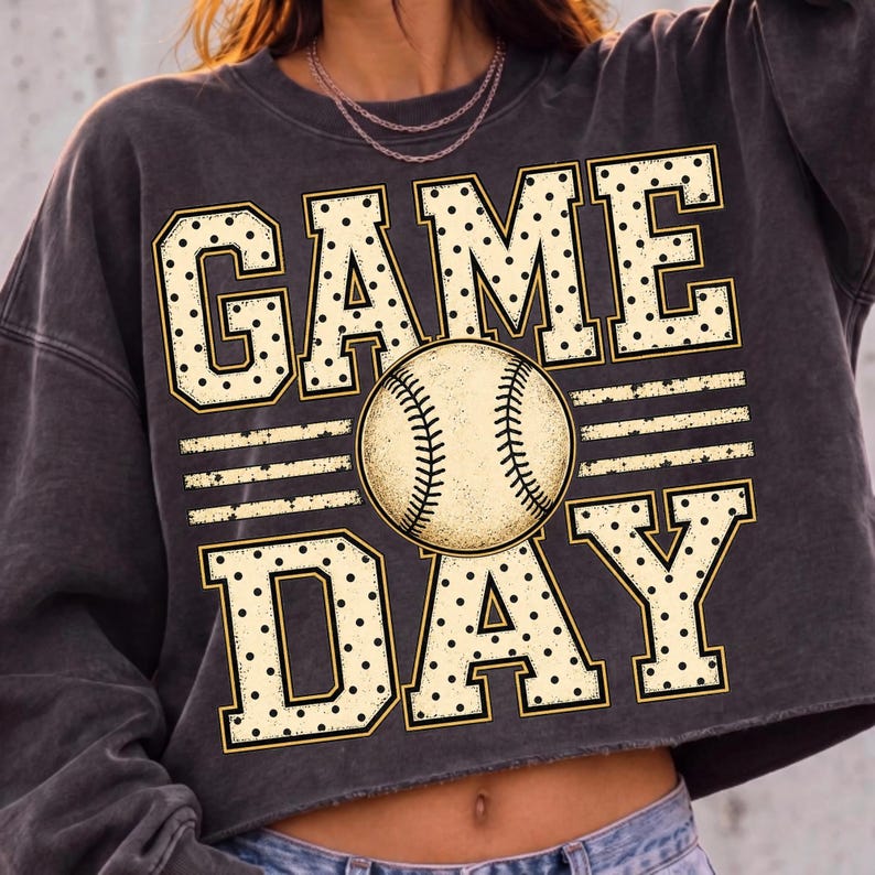 May include: A charcoal grey cropped sweatshirt featuring a graphic print with the text GAME DAY in large, cream-coloured block letters with black polka dots. A central baseball illustration with visible stitching sits between the two words.
