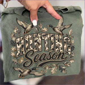 May include: An olive green t-shirt featuring a graphic print with the text "HUNTING SEASON" in a camouflage pattern. The design includes three flying mallard ducks and a border of oak leaves surrounding the bold, block-style lettering.