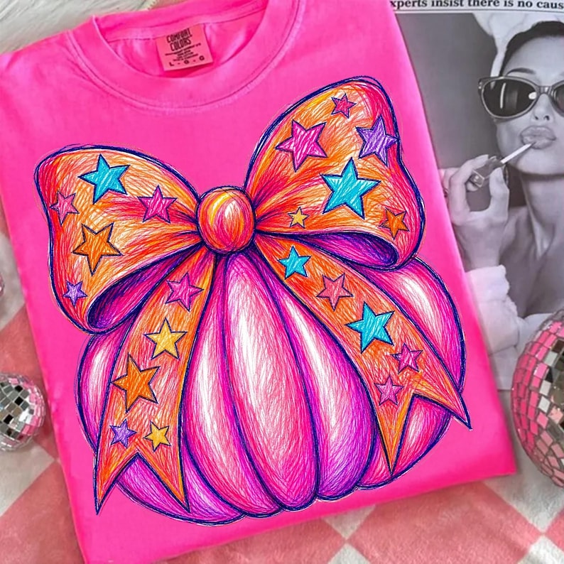 May include: A bright pink t-shirt featuring a large, hand-drawn style graphic of a pumpkin decorated with a prominent bow. The pumpkin and bow are filled with vibrant scribbled lines in shades of orange, purple, and pink, accented by numerous colorful stars in blue, yellow, and pink.