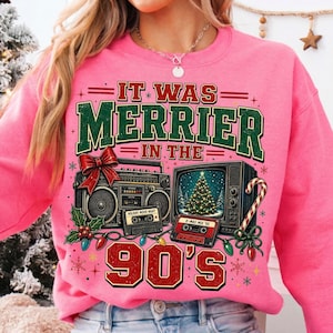 Może przedstawiać: Jaskrawor&oacute;żowa bluza z grafiką i napisem IT WAS MERRIER IN THE 90'S. Wz&oacute;r przedstawia czarny radiomagnetofon, stary telewizor z choinką, kasety magnetofonowe, czerwoną kokardę oraz świąteczne lampki.