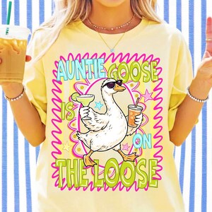 K&ouml;nnte beinhalten: Gelbes T-Shirt mit einer Grafik einer Gans mit Sonnenbrille, die einen Cocktail und ein Getr&auml;nk h&auml;lt. Der Text "Auntie Goose is on the Loose" ist in einem farbenfrohen Design dargestellt.