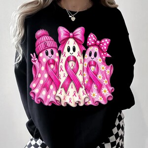 Puede incluir: Una sudadera negra de manga larga con un gr&aacute;fico de tres fantasmas decorados con lazos rosas de concienciaci&oacute;n sobre el c&aacute;ncer de mama. Los fantasmas llevan lunares rosas, margaritas y lazos, y uno de ellos lleva un gorro de punto rosa.