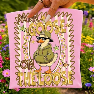 May include: A light pink fabric item featuring a graphic of a goose wearing a green hoodie, a baseball cap, and pixelated sunglasses. The text DADA GOOSE ON THE LOOSE is printed in gold and brown letters, surrounded by a decorative oval and stars.