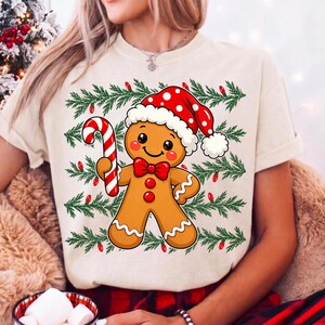 Puede incluir: Una camiseta de color crema con un gr&aacute;fico de un hombre de jengibre que lleva un gorro de Pap&aacute; Noel rojo con lunares blancos y una pajarita roja. El hombre de jengibre sostiene un bast&oacute;n de caramelo rojo y blanco sobre ramas de pino verdes.