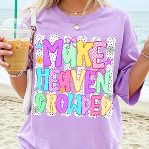 May include: A light purple t-shirt featuring a graphic print with the text "MAKE HEAVEN CROWDED" in colorful, patterned block letters. The design includes black and white polka dot vertical stripes, small flowers, and pink ribbon accents.