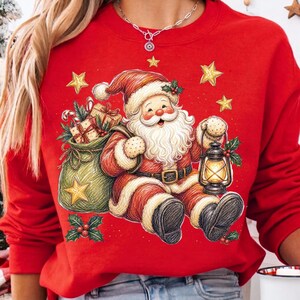 K&ouml;nnte beinhalten: Ein leuchtend rotes Sweatshirt mit Rundhalsausschnitt und einer festlichen Grafik des Weihnachtsmanns mit wei&szlig;em Bart, rotem Anzug, einer leuchtenden Laterne und einem gr&uuml;nen Sack voller Geschenke, umgeben von gelben Sternen und Stechpalmen.
