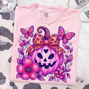 K&ouml;nnte beinhalten: Ein hellrosa T-Shirt mit einer Grafik eines K&uuml;rbisgesichts mit herzf&ouml;rmiger Nase, das mit einer Schleife im Leopardenmuster verziert ist. Das Design zeigt leuchtend rosa Blumen, Beeren und zwei Schmetterlinge in Rosa- und Violettt&ouml;nen.
