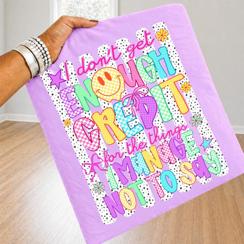 Pode incluir: Um artigo de tecido roxo claro com uma estampa gr&aacute;fica com o texto 'I don't get ENOUGH CREDIT for the things I MANAGE NOT TO SAY'. O design inclui um rosto sorridente amarelo, padr&otilde;es xadrez coloridos, pequenas flores e estrelas.